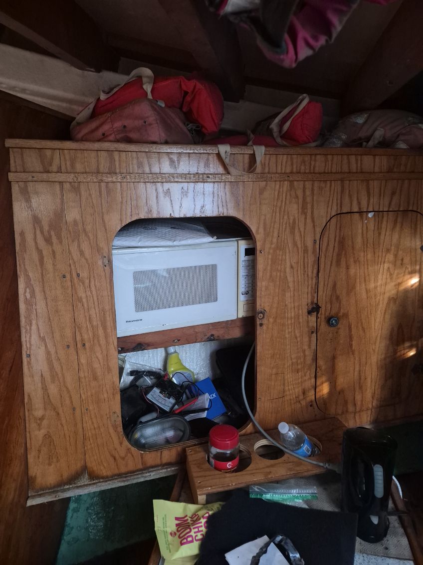A vintage white microwave oven tucked into a wooden storage cabinet on a boat or camper interior, with various household items and bags visible around it.
