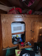 A vintage white microwave oven tucked into a wooden storage cabinet on a boat or camper interior, with various household items and bags visible around it.