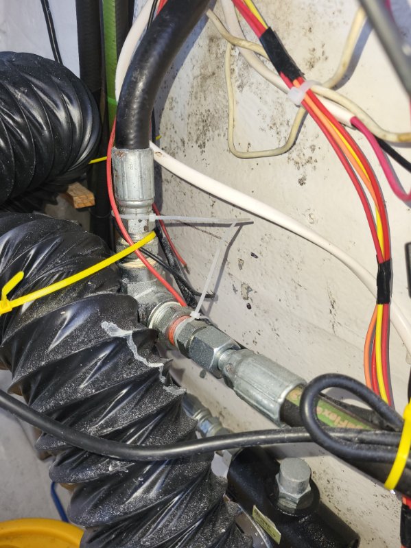 Close-up of industrial piping and wiring in a mechanical or utility setting, featuring a corrugated black hose, metal couplings, and multicolored wires against a dirty concrete wall.