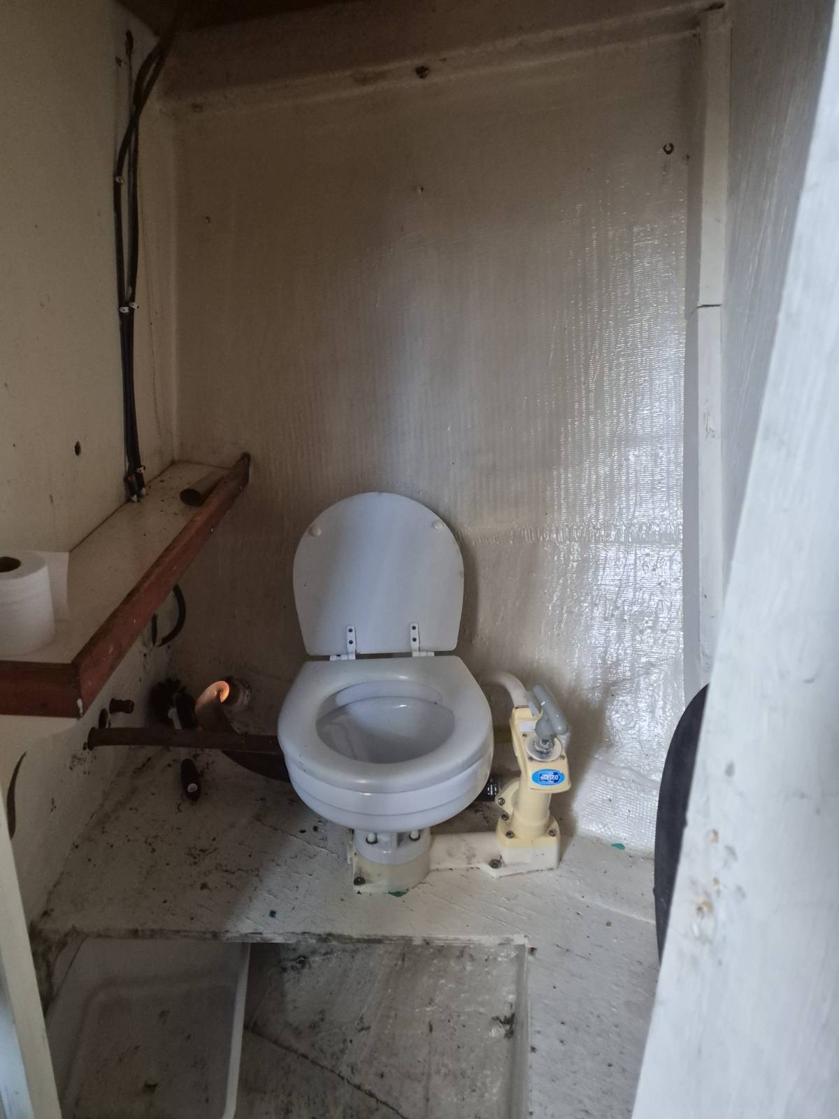 A compact, utilitarian toilet stall with a white ceramic toilet, open lid, and visible plumbing fixtures against a textured beige wall.