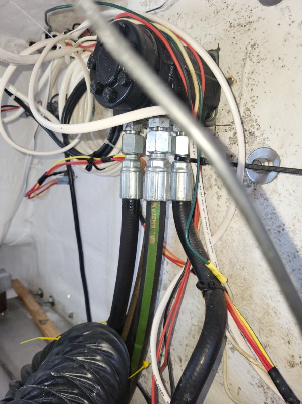 Close-up of an electrical or plumbing junction with multiple black, white, red, yellow, and green wires, three silver metal connector fittings, black hoses, and a corrugated black pipe sleeve against a dirty off-white wall.