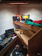 A cluttered wooden bunk bed or storage area with folded textiles, tools, and equipment on a shelf and floor, photographed in a real interior space.