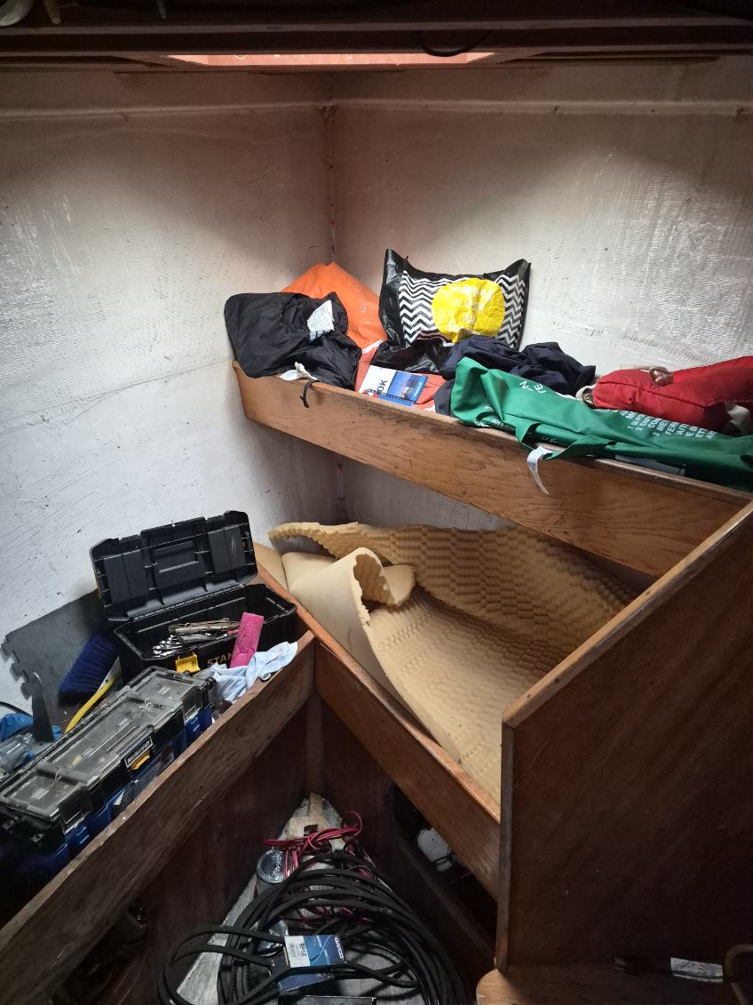 A cluttered wooden bunk bed or storage area with folded textiles, tools, and equipment on a shelf and floor, photographed in a real interior space.