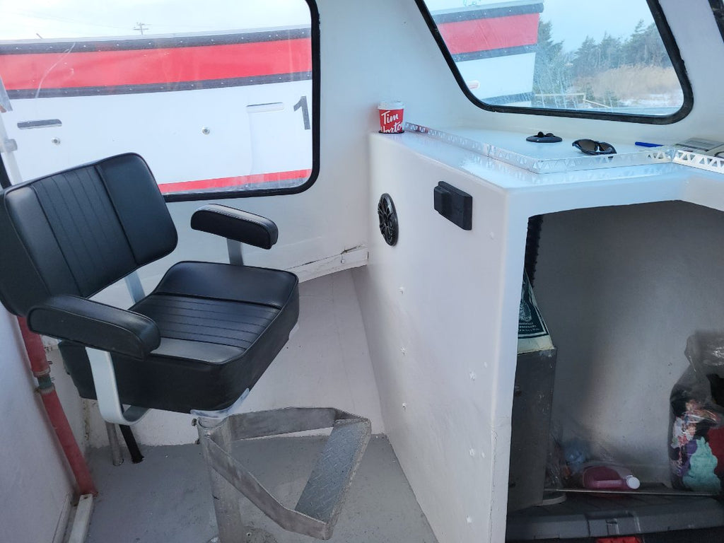 A black vinyl boat helm seat with armrests, positioned next to a white boat cabin console with windows and a small storage compartment.
