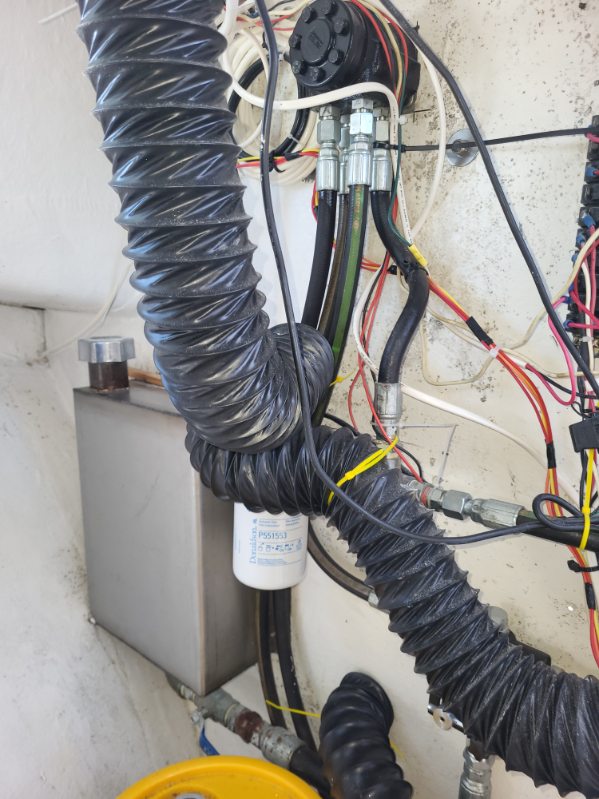 Close-up of a mechanical setup with black corrugated flexible hoses, bundled wires, a white cylindrical filter, a gray metal box, and a concrete wall background.
