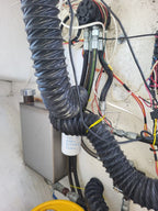 Close-up of a mechanical setup with black corrugated flexible hoses, bundled wires, a white cylindrical filter, a gray metal box, and a concrete wall background.