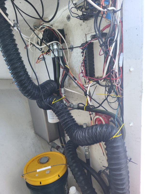A close-up view of an electrical or mechanical compartment with numerous black corrugated hoses, bundles of red, white, and black wires, metal conduit, and a yellow-and-black bucket at the bottom left.