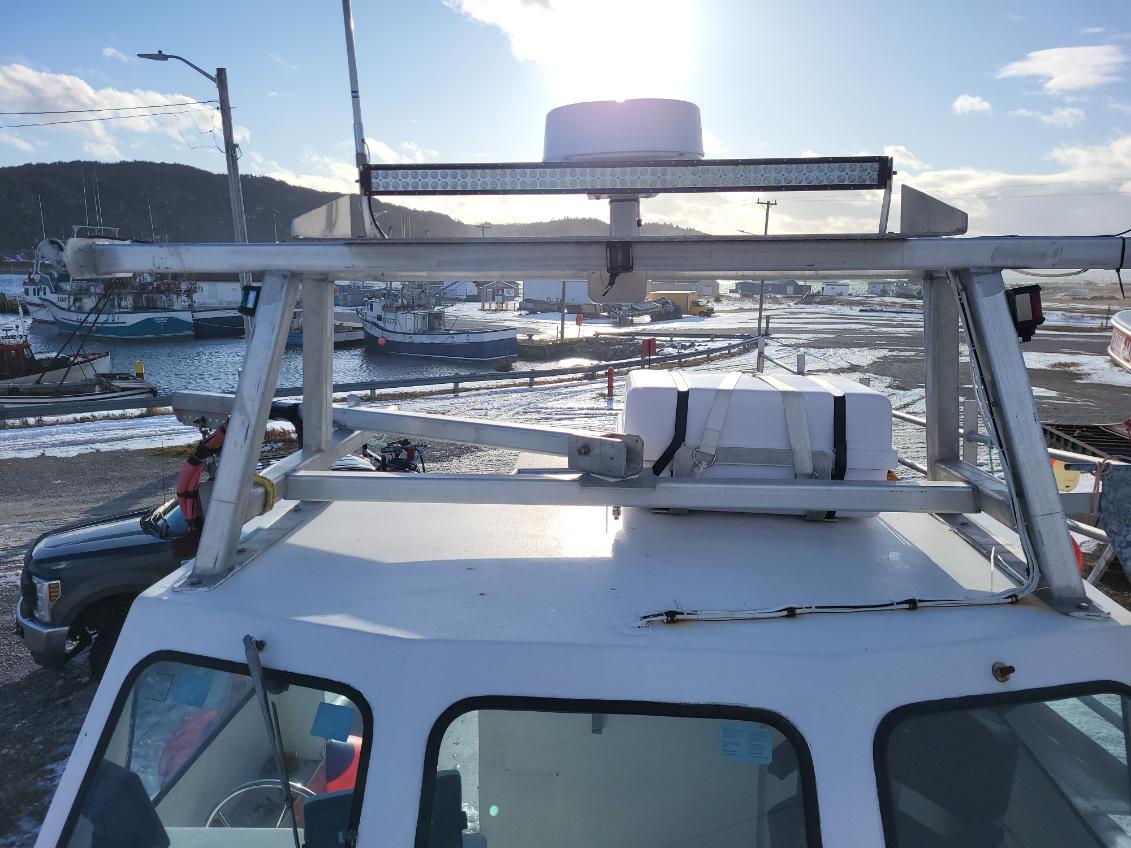 A white boat cabin roof with a metal mounting frame and a long LED light bar mounted above, photographed outdoors at a harbor with boats, water, and shoreline in the background.