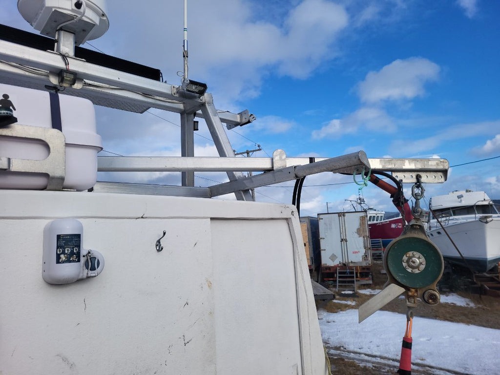 A close-up view of a white utility or boat-related vehicle or equipment unit outdoors under a blue sky with clouds, showing metal mounting hardware, a green circular component, and other industrial equipment in the background.