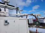 A close-up view of a white utility or boat-related vehicle or equipment unit outdoors under a blue sky with clouds, showing metal mounting hardware, a green circular component, and other industrial equipment in the background.