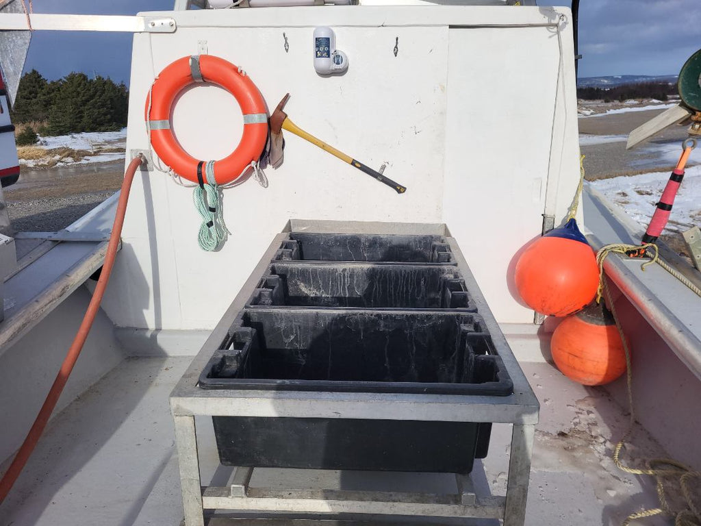 A black rectangular fish holding box on a metal frame, set on the deck of a white boat with orange buoys and a life preserver visible in the background.
