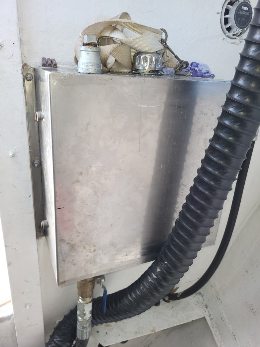 A stainless steel marine utility box mounted on a white wall, with a beige strap and metal fittings on top, a black corrugated hose running down the right side, and a metal pipe connection at the bottom.
