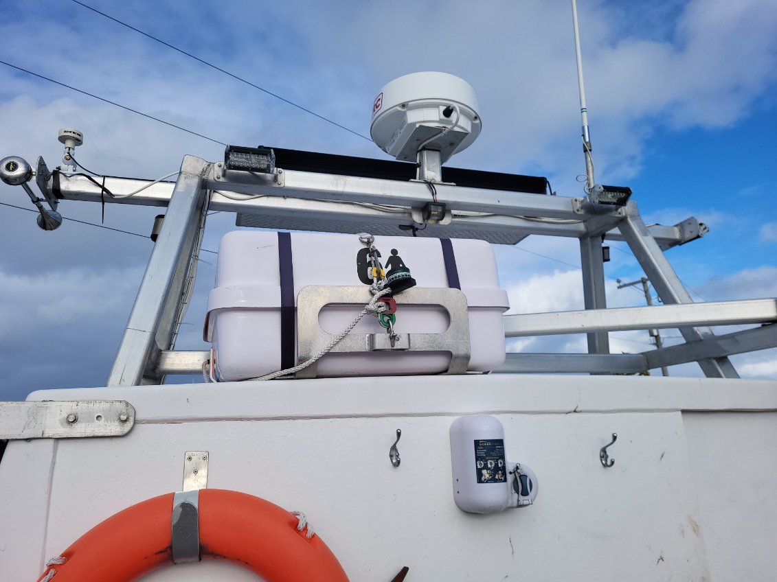 A close-up view of a white boat or vessel deck with a metal mounting frame, a white radar or communication antenna dome, a small bell, and an orange life preserver against a blue sky with clouds.