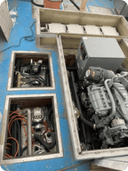 An open mechanical engine compartment with exposed metal frames, hoses, wiring, and a large gray engine block on a blue surface.
