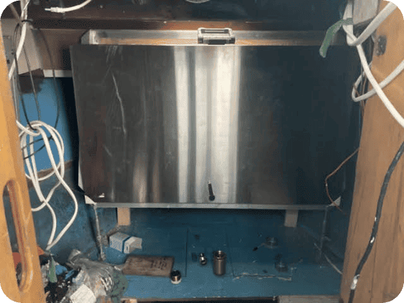 A large rectangular stainless steel panel or door mounted inside a blue-framed opening, surrounded by visible wiring, tools, and workshop materials.