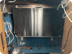 A large rectangular stainless steel panel or door mounted inside a blue-framed opening, surrounded by visible wiring, tools, and workshop materials.