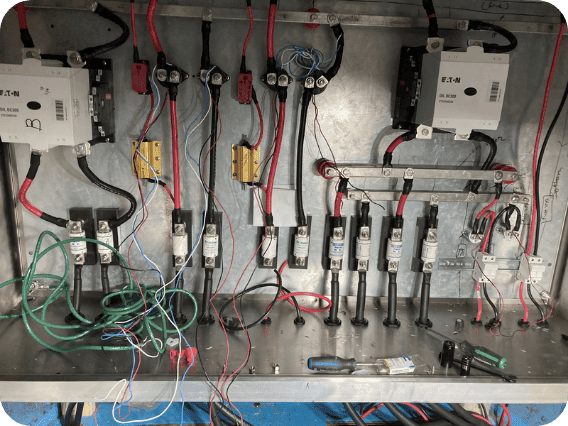 An electrical panel or control cabinet interior with multiple circuit breakers, fuses, wires, terminal blocks, and metal components arranged on a metal chassis.