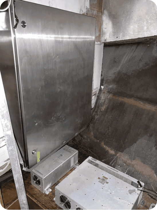 A large brushed stainless steel appliance panel or cabinet shown in a rough industrial or warehouse setting, with two metal electrical or control boxes on the floor nearby.