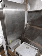 A large brushed stainless steel appliance panel or cabinet shown in a rough industrial or warehouse setting, with two metal electrical or control boxes on the floor nearby.