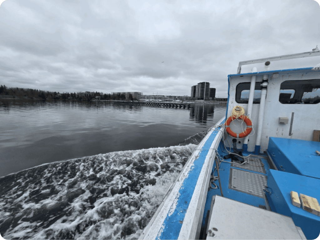 A blue and white boat moving across calm water under an overcast sky, with a life preserver on the side and a distant shoreline with trees and buildings.