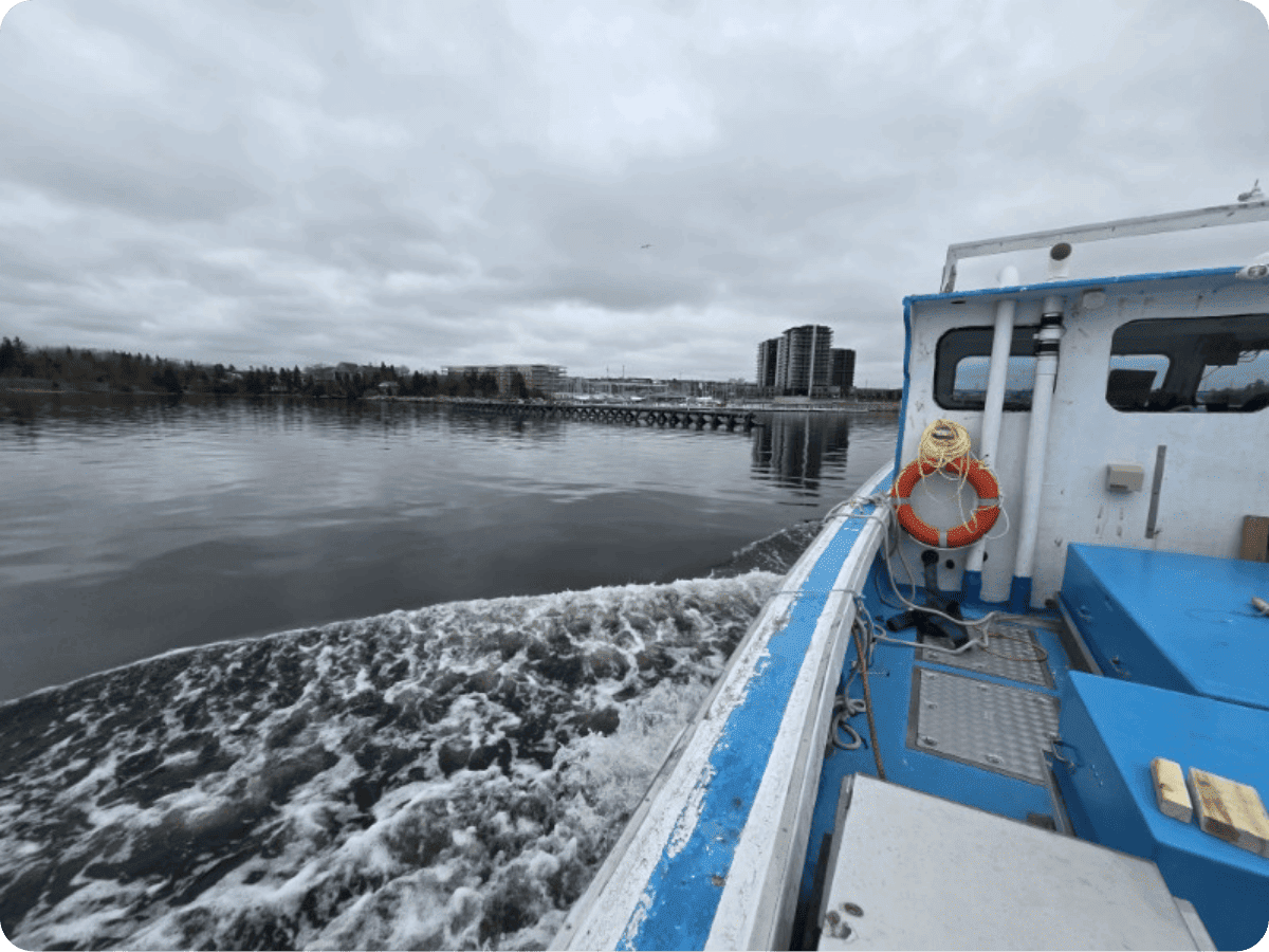 A blue and white boat moving across calm water under an overcast sky, with a life preserver on the side and a distant shoreline with trees and buildings.