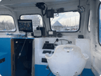 The helm station of a boat cabin featuring a white molded seat, navigation screens, a steering wheel, and various electronic equipment mounted on the white and blue painted interior walls.