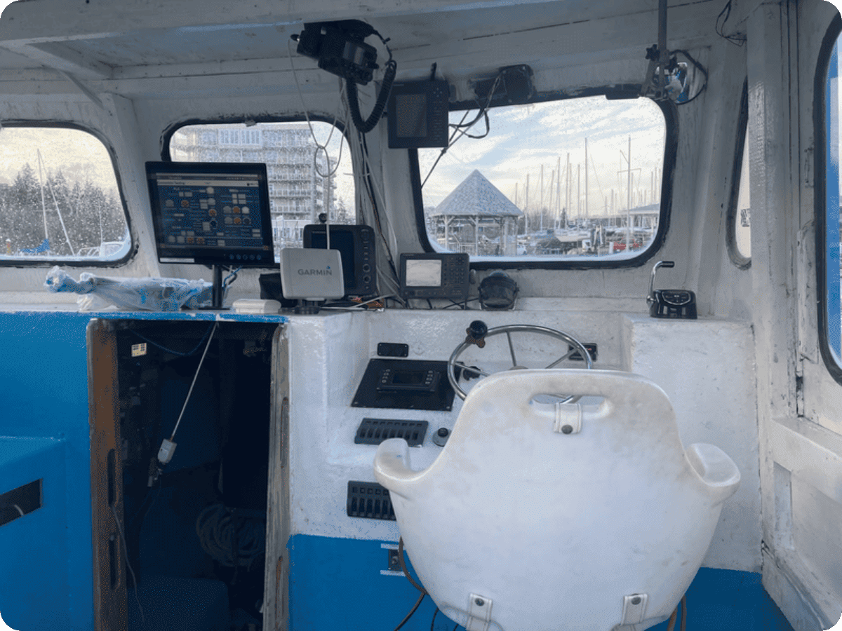 The helm station of a boat cabin featuring a white molded seat, navigation screens, a steering wheel, and various electronic equipment mounted on the white and blue painted interior walls.