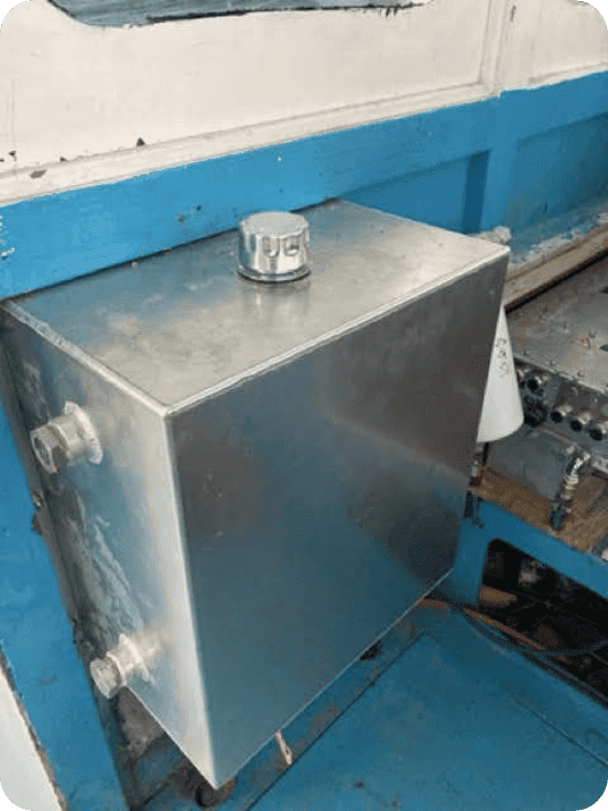 A rectangular stainless steel tank with a silver cap on top and two side fittings, mounted against a blue and white painted structure.