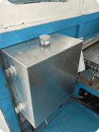 A rectangular stainless steel tank with a silver cap on top and two side fittings, mounted against a blue and white painted structure.