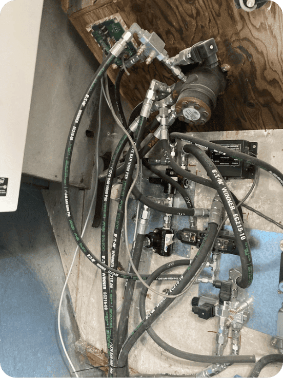 Close-up of a mechanical hydraulic or pneumatic control assembly with multiple black hoses, metal fittings, a small motor or pump component, and an electronic control module mounted against a wood and metal panel.